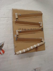 A large rectangular piece of cardboard taped to a white wall with smaller strips of cardboard taped in a zig-zag pattern to create a marble run