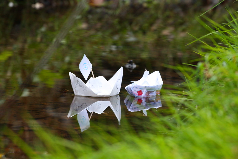 Two white paper origami boats decorated with kids' drawings and toothpick masts with paper sails floating in a small stream lined with green grasses