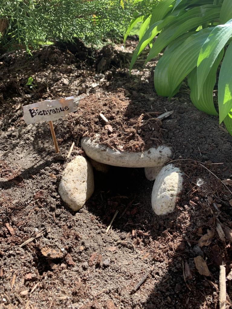 A shallow cave /den made of river rocks and mulch in a garden. Small welcome sign made of birch bark on the outside of the structure.