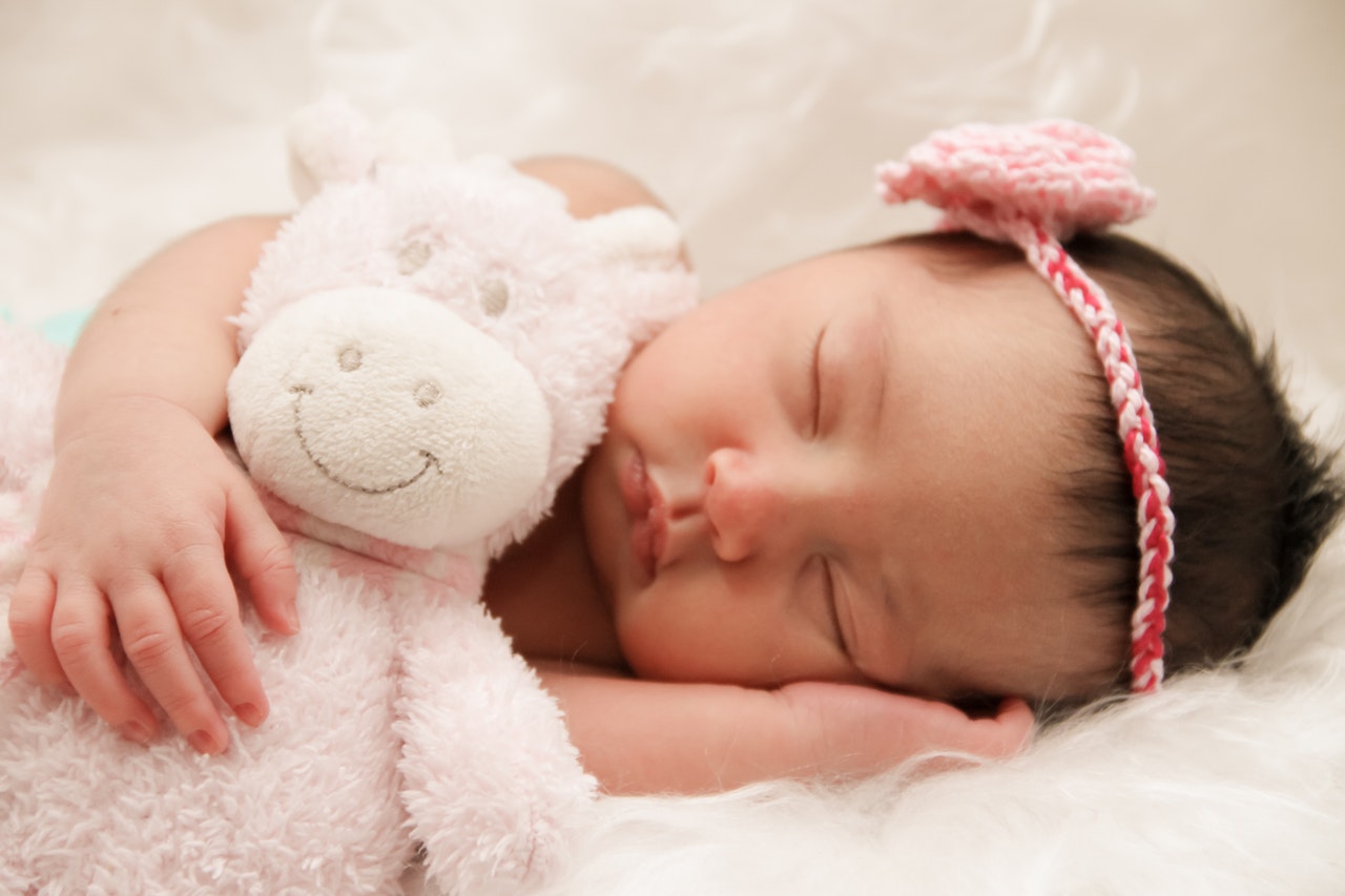 Baby sleeping on her side with a pink ribbon and crocheted flower on her head, and holding a stuffed, white bear.
