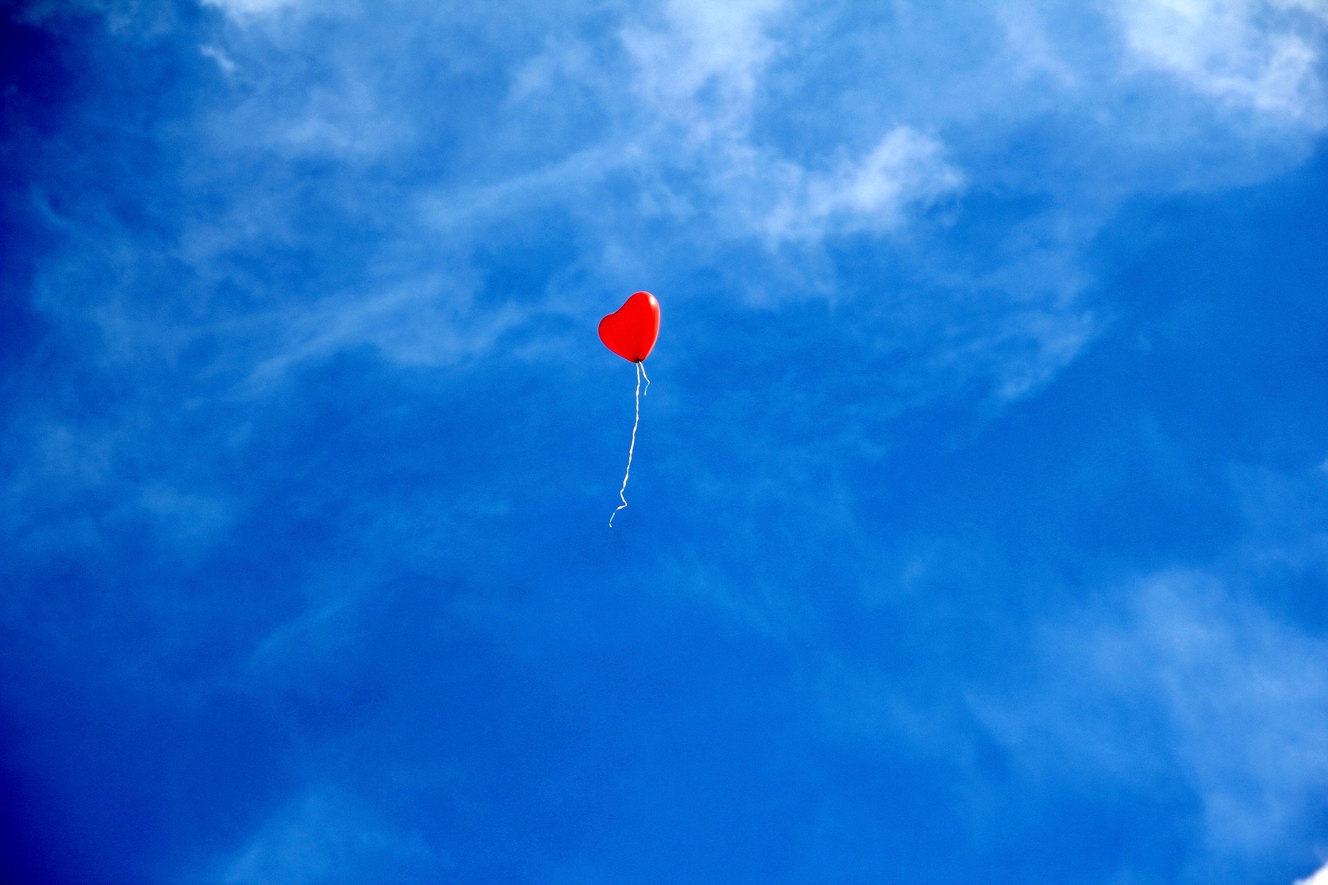 Heart shaped red balloon high in the sky with a white string hanging down. The sky is a vibrant blue with whispy clouds around.
