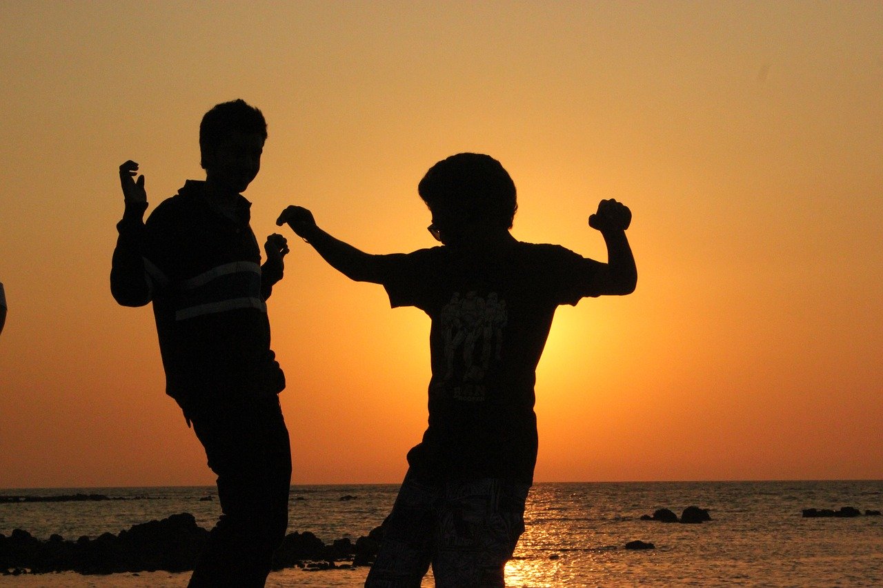 Silhouette of two children dancing on a beach with an orange sunset behind them.