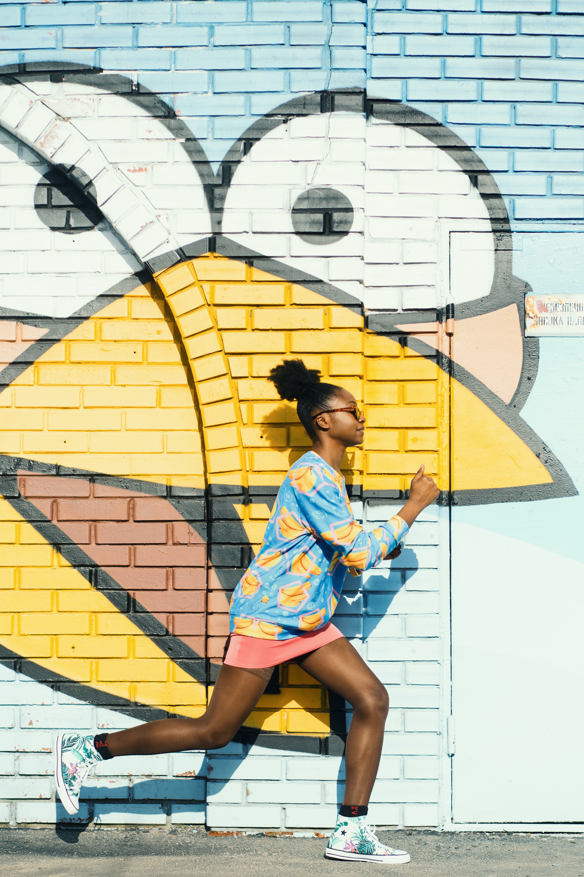 Girl jogging in front of brick wall that has a painted picture of a bird face with a yellow beak and white bulging eyes.
