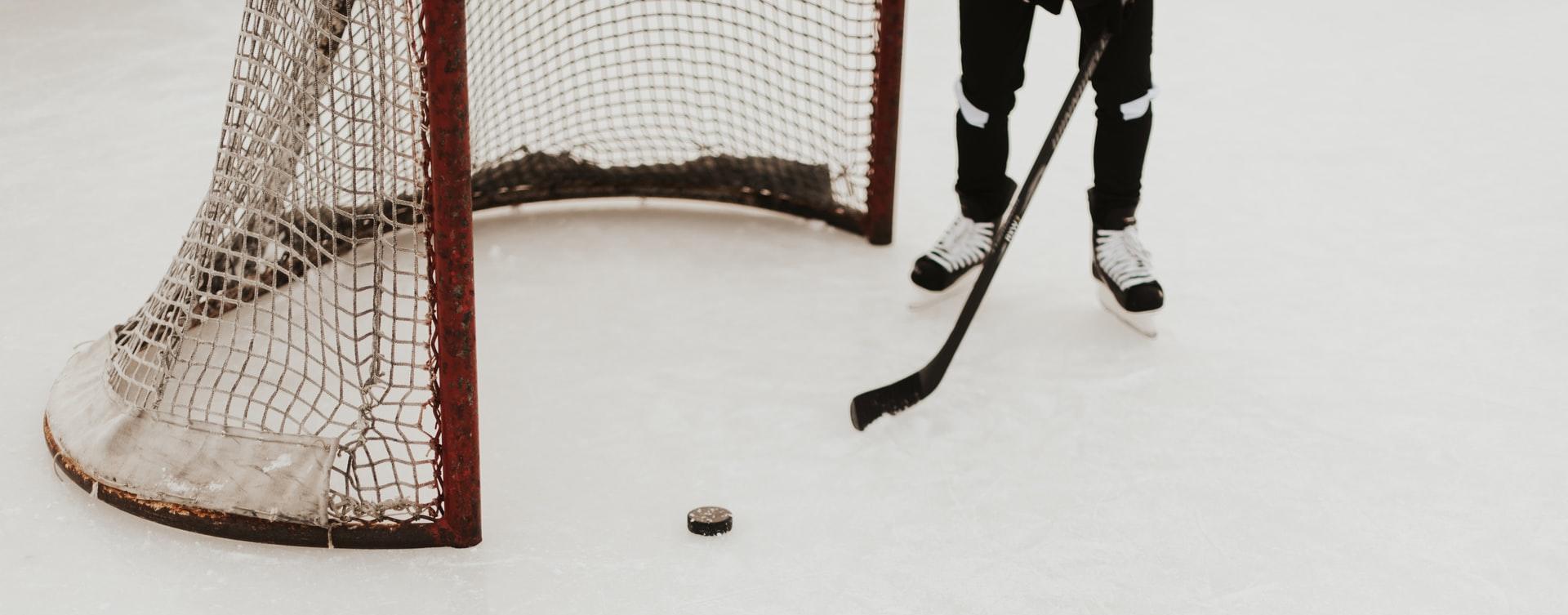 A hockey net on an ice rink, with a puck in front of the net and a person on skates holding a stick, but you can only see the person's legs and the stick.