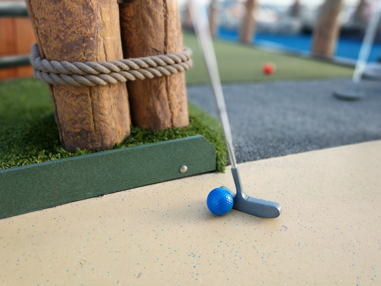 A blue golf ball and metal golf putter leaning up against a 2 wooden posts that are tied together with rope, anchored in green artificial turf.